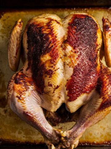 A top-down view of a partially cooked rotisserie chicken on a roasting tray, with crispy golden-brown skin developing, surrounded by rendered juices and seasoning.