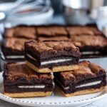 A close-up shot of four freshly baked slutty brownies stacked on a white plate, showing distinct layers of golden cookie base, Oreo center, and fudgy brownie top, with a baking pan in the background.