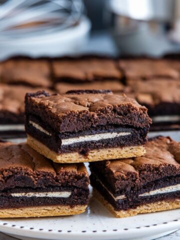 A close-up shot of four freshly baked slutty brownies stacked on a white plate, showing distinct layers of golden cookie base, Oreo center, and fudgy brownie top, with a baking pan in the background.