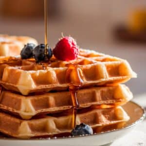 A stack of golden-brown Krusteaz waffles with crisp edges, topped with fresh berries and drizzled with maple syrup. Warm morning light highlights their fluffy texture