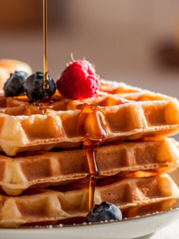 A stack of golden-brown Krusteaz waffles with crisp edges, topped with fresh berries and drizzled with maple syrup. Warm morning light highlights their fluffy texture