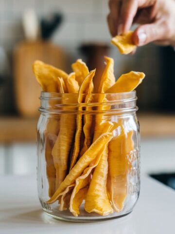 Homemade dehydrated mango slices in a glass jar on a white kitchen counter, with a hand reaching for a piece, and blurred kitchen items in the background.