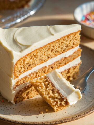 A moist, homemade gluten-free vanilla cake Slice with creamy white frosting, crumbs on a white plate, and a fork holding a bite.