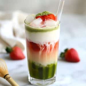 An Iced strawberry matcha latte in a clear glass, showing layered strawberries, milk, and matcha with a glass straw.