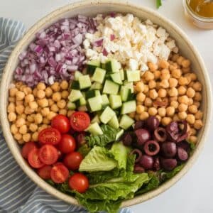 top-down view of a Mediterranean chopped salad in a ceramic bowl, neatly arranged before mixing. A jar of vinaigrette and parsley leaves sit on a white kitchen counter.