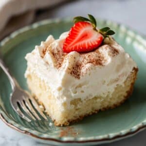 A moist slice of homemade tres leches cake on a pink fluted plate, topped with whipped cream, cinnamon dusting, and a sliced strawberry. A fork rests beside the cake, with milk pooling slightly at the base.