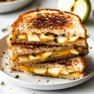 Stacked apple grilled cheese halves on a white plate with melted cheese, apple slices, and golden crust. Homemade look on rustic table with natural light.