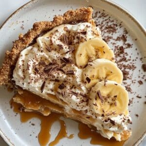 A homemade slice of Banoffee Pie with caramel and bananas, topped with whipped cream and chocolate shavings on a white plate.