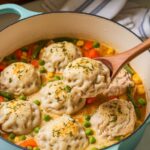 A homemade pot of Bisquick chicken and dumplings with fluffy herb-speckled dumplings in a rich, golden broth, filled with chicken and vegetables. A spoon lifts one dumpling, showing its soft texture.