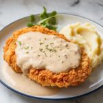 A crispy, golden chicken fried steak topped with creamy pepper gravy, served with mashed potatoes on a white plate. Homemade and slightly rustic.