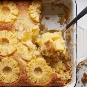 A golden, crispy pineapple dump cake in a white baking dish with a spoonful removed, showing juicy pineapple filling underneath.