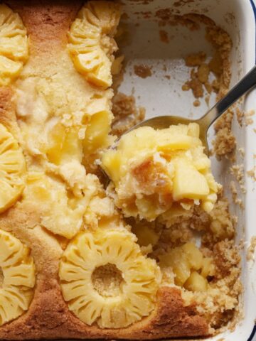 A golden, crispy pineapple dump cake in a white baking dish with a spoonful removed, showing juicy pineapple filling underneath.