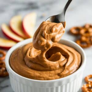 A spoon lifting a thick scoop of peanut butter dip from a bowl, with apple slices and pretzels nearby.