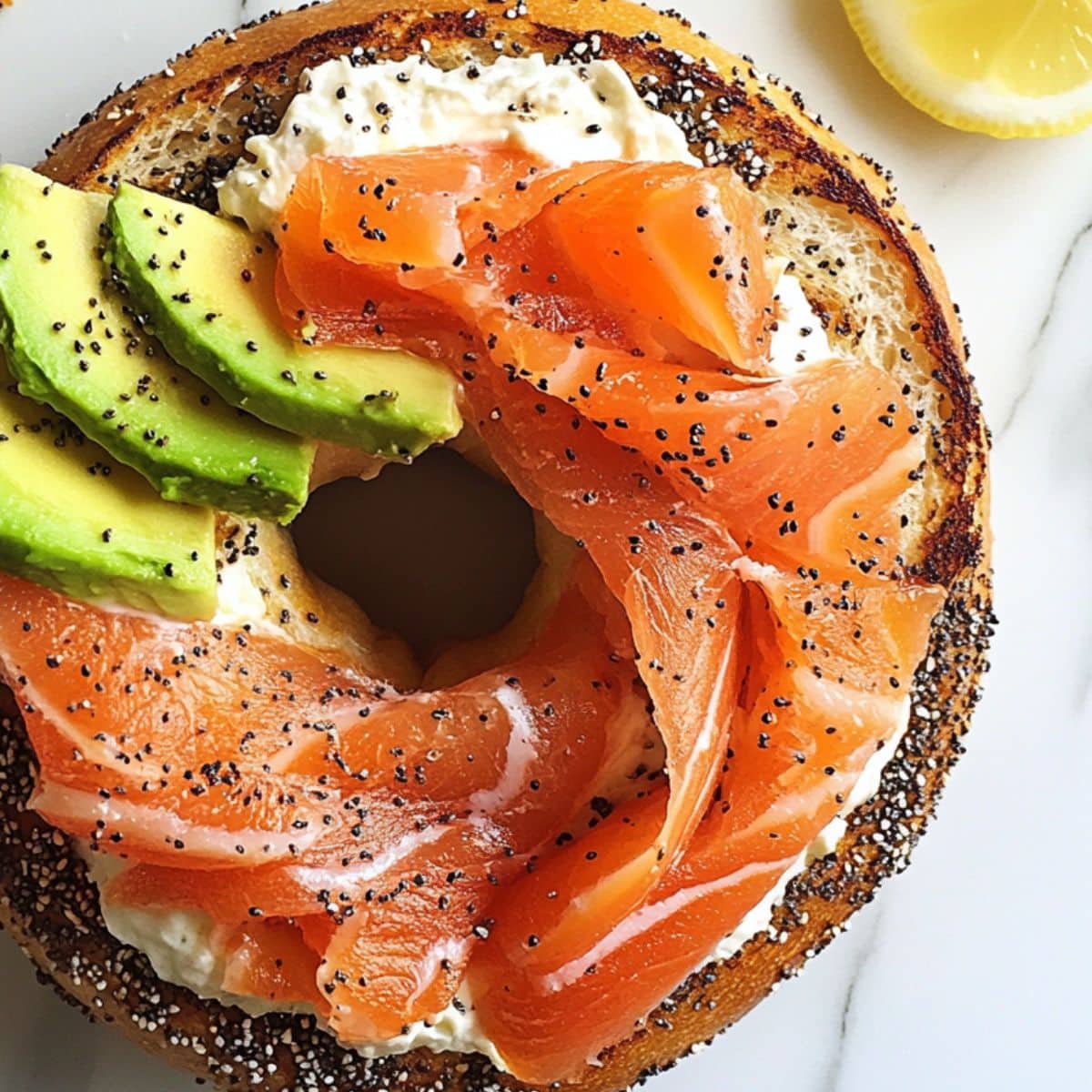 Cream cheese-covered bagel topped with folded smoked salmon and avocado slices, beginning the smoked salmon breakfast build, on a white marble counter.