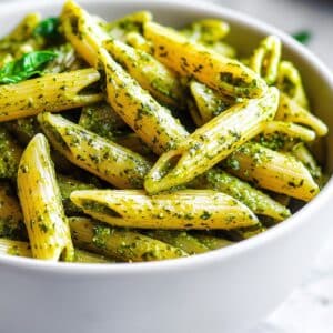 Overhead close-up of juicy penne pasta coated in vibrant green pesto, topped with fresh basil. Homemade, real texture on a white counter.