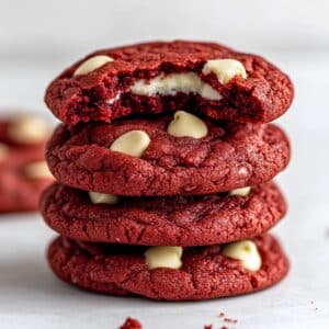 Stack of red velvet cookies with white chocolate chips, top cookie bitten to reveal moist red center. Homemade look with natural lighting and visible crumbs.