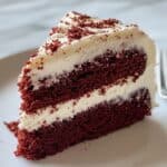 Slice of red velvet cake with two layers of fluffy cream cheese frosting, topped with red cake crumbs on a white plate. Soft, moist texture and vibrant color make it a classic dessert favorite.