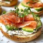 Smoked salmon breakfast bagel with cream cheese, avocado slices, and fresh toppings on a white marble kitchen counter. A homemade smoked salmon breakfast perfect for weekends.