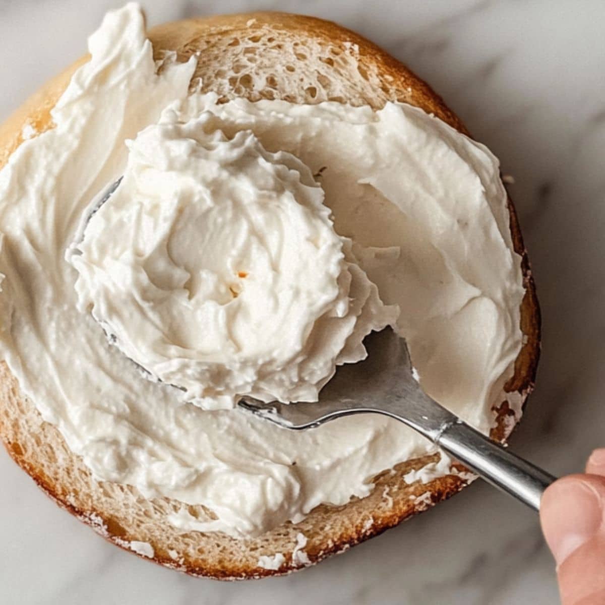 Toasted bagel spread generously with whipped cream cheese, forming the base for a smoked salmon breakfast, photographed casually on a white marble counter.