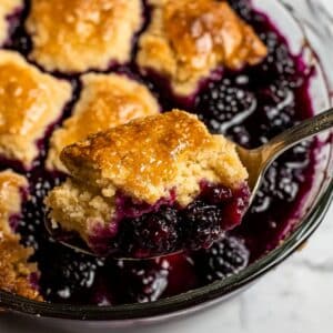 Golden-topped baked blackberry cobbler recipe with juicy berries scooped from a glass baking dish, showing a moist biscuit topping and bubbling berry filling.