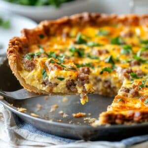 Golden breakfast quiche with flaky crust, sausage, and cheddar. A slice is lifted to reveal a fluffy, creamy interior with fresh herbs on top.