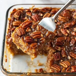 Slice of homemade pecan pie dump cake lifted from a glass dish. Gooey center and golden pecans on top, real kitchen setting.