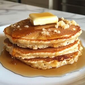Stack of homemade sourdough discard pancakes with syrup and butter on white counter. Fluffy, golden, imperfectly shaped pancakes from overhead.