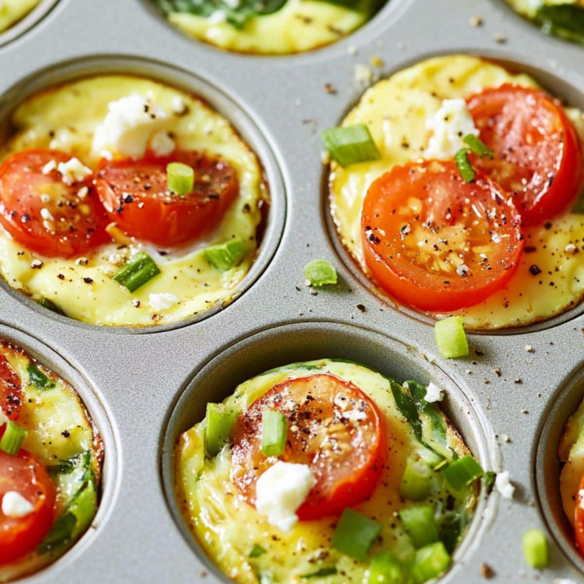 Close-up of unbaked egg muffins with tomatoes, spinach, and feta in a muffin tin. Sprinkled with black pepper and green onions.