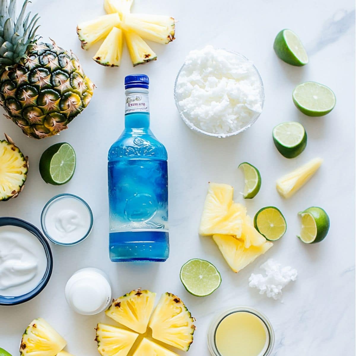 Flat lay of real Blue Hawaiian Margarita ingredients on white counter, including tequila, blue curaçao, lime, and pineapple juice.