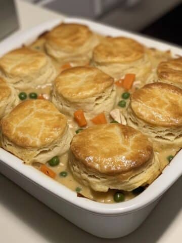 Golden baked biscuit topped chicken pot pie casserole in a white ceramic dish. Creamy filling with visible chicken, carrots, and peas.