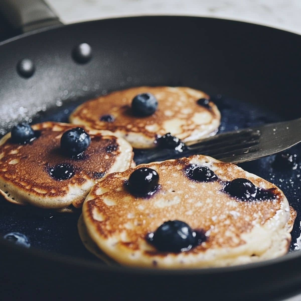 Blueberry pancakes cooking on a skillet with golden edges and popping berries.