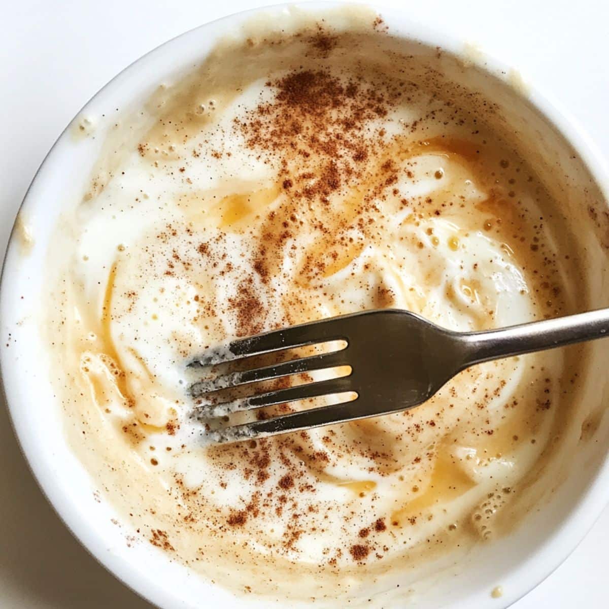 Homemade egg custard with cinnamon, vanilla, and sugar. Whisk resting in bowl on white counter with light spills.