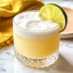 Tequila sour with thick white foam and lime wheel garnish in a clear rocks glass.
