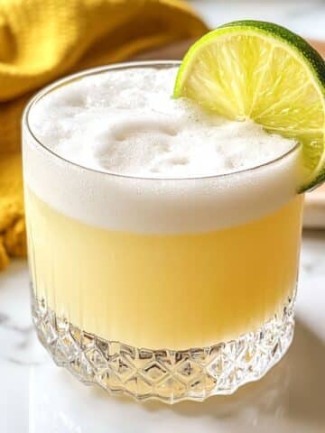 Tequila sour with thick white foam and lime wheel garnish in a clear rocks glass.