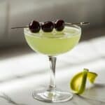 Homemade Last Word cocktail in a coupe glass with cherry garnish and lime twist on white counter, showing natural shadows and pale green color.