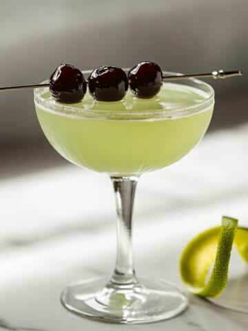 Homemade Last Word cocktail in a coupe glass with cherry garnish and lime twist on white counter, showing natural shadows and pale green color.