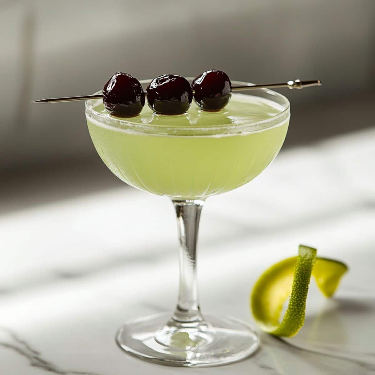 Homemade Last Word cocktail in a coupe glass with cherry garnish and lime twist on white counter, showing natural shadows and pale green color.