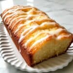 Lemon pound cake sliced on a white plate, topped with a light lemon glaze. Moist crumb and real kitchen texture with natural light.