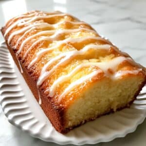 Lemon pound cake sliced on a white plate, topped with a light lemon glaze. Moist crumb and real kitchen texture with natural light.