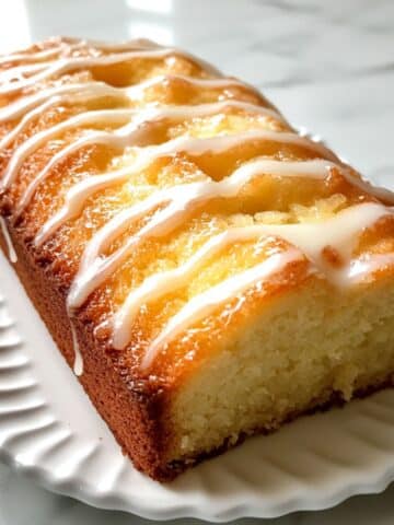Lemon pound cake sliced on a white plate, topped with a light lemon glaze. Moist crumb and real kitchen texture with natural light.