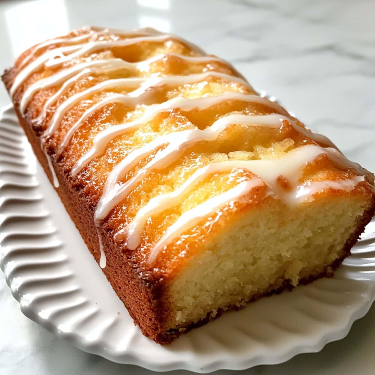Lemon pound cake sliced on a white plate, topped with a light lemon glaze. Moist crumb and real kitchen texture with natural light.
