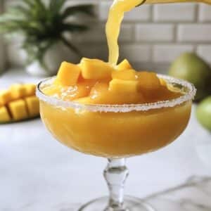Pouring thick mango margarita into a Tajín-rimmed glass, topped with fresh mango cubes and wedge.
