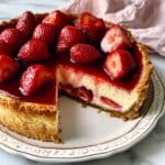 Sliced roasted strawberry cheesecake topped with glossy jam and fresh strawberries, served on a white plate with a golden graham crust.