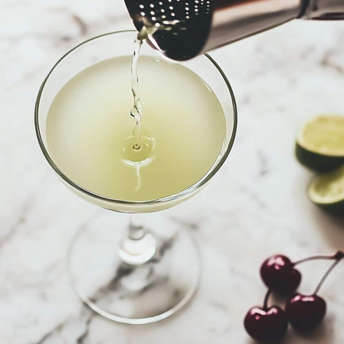 Pouring pale green Last word cocktail through a strainer into a coupe glass with cherry and lime garnish ready.