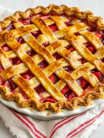 Close-up of a golden, lattice-topped strawberry rhubarb pie with bubbling fruit filling, rustic crust, and juicy homemade texture on white towel and wooden counter.