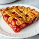 Golden lattice-topped strawberry rhubarb pie slice on a white plate, showing juicy strawberries and glossy filling inside a flaky crust.