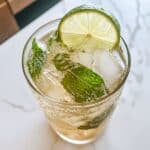 Close-up of virgin mojito with mint, lime slice, and ice cubes in a glass. Fizzy bubbles and fresh herbs on a white marble kitchen counter.