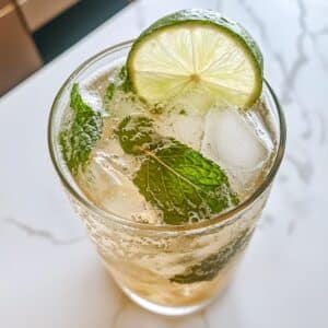 Close-up of virgin mojito with mint, lime slice, and ice cubes in a glass. Fizzy bubbles and fresh herbs on a white marble kitchen counter.