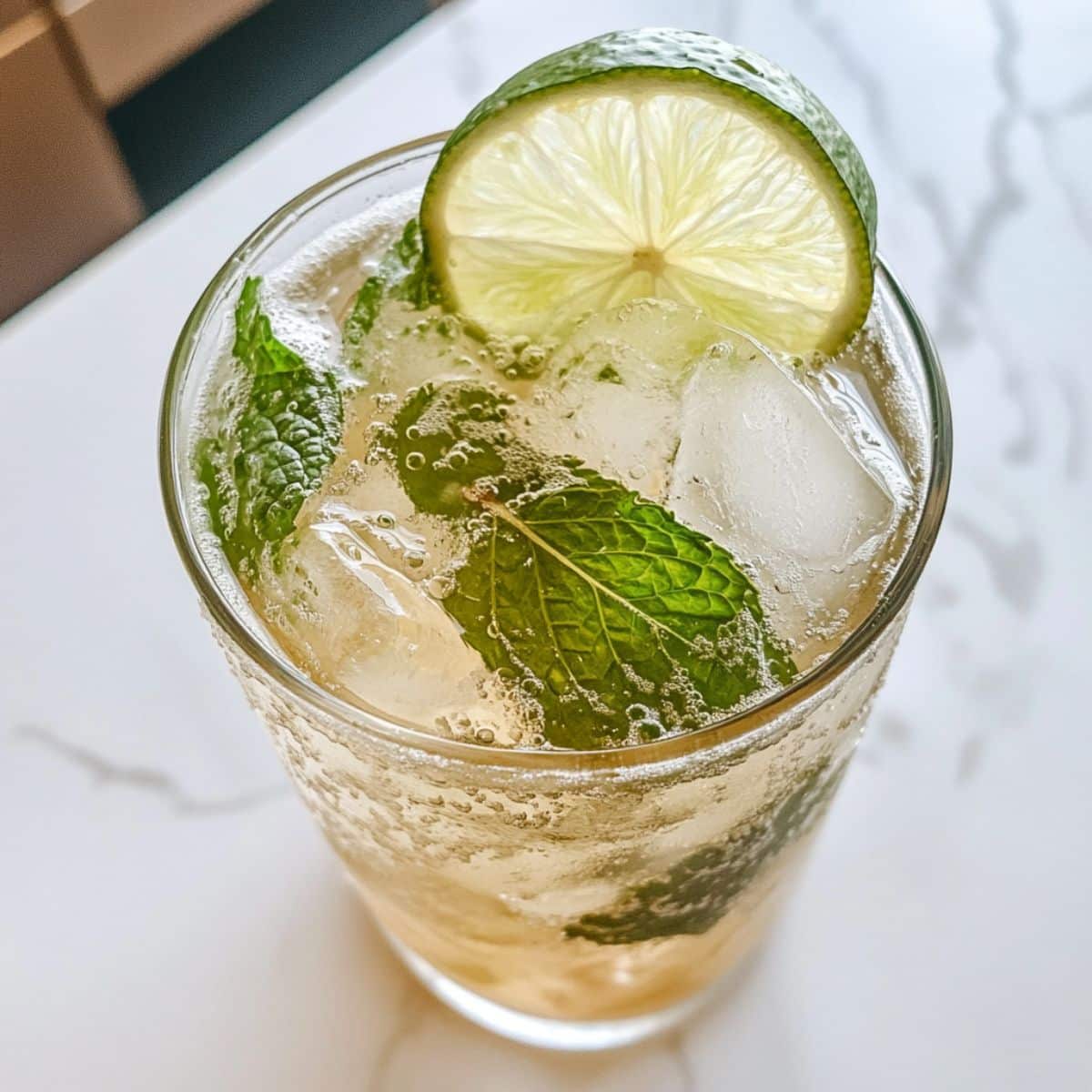 Close-up of virgin mojito with mint, lime slice, and ice cubes in a glass. Fizzy bubbles and fresh herbs on a white marble kitchen counter.