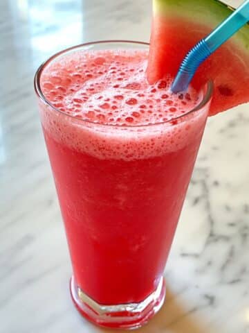 Tall glass of homemade watermelon juice with frothy top, blue straw, and watermelon wedge.